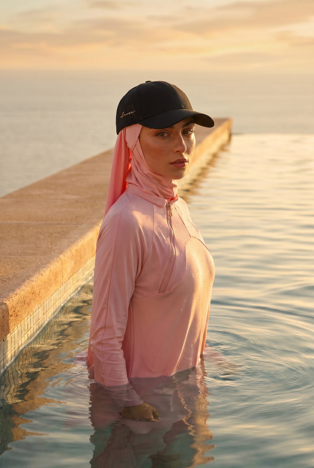 Model trägt die schwarze MIKA Cap mit goldenem Logo-Stick über dem Swim-Hijab – am Wasser im Sonnenuntergang