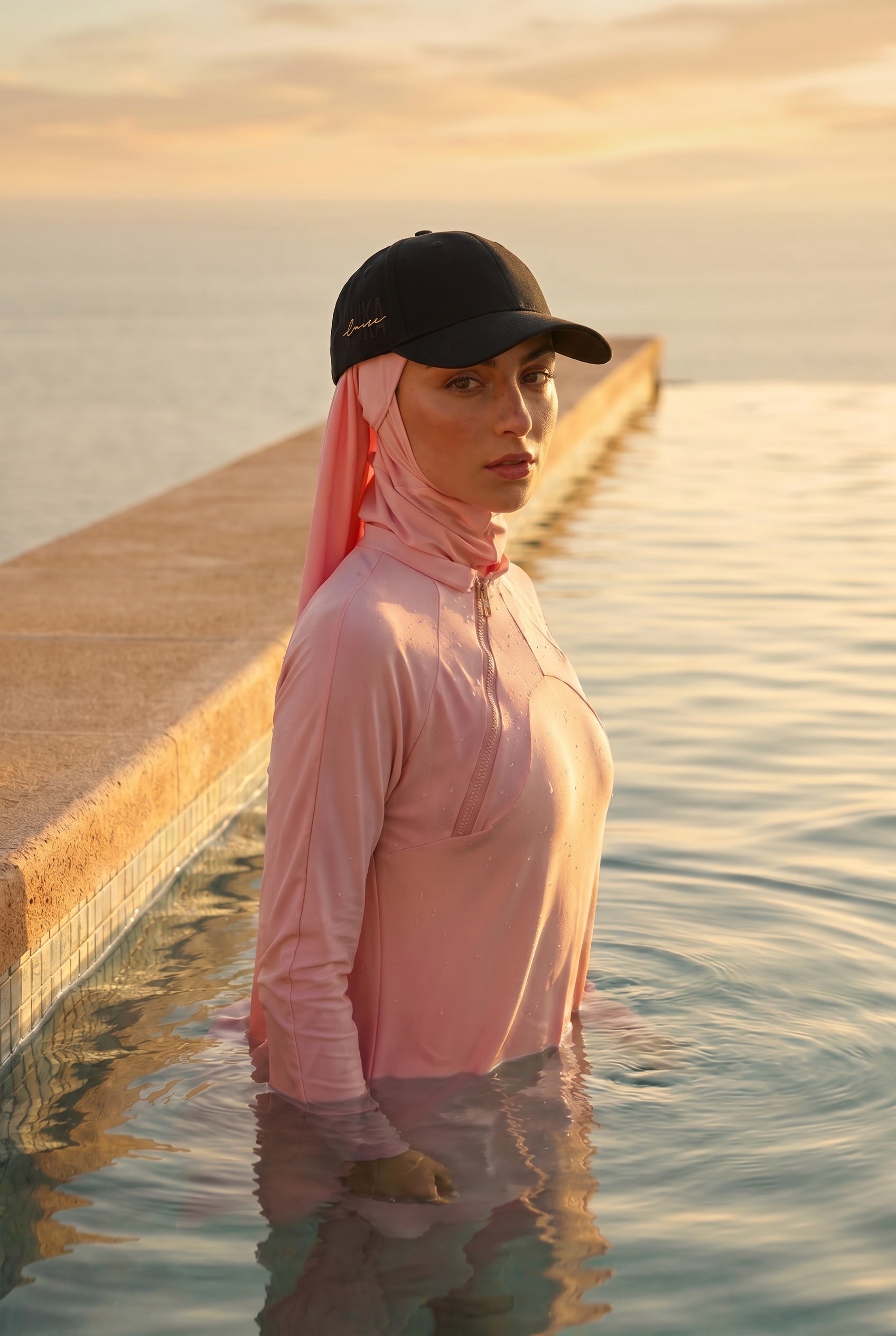 Model trägt die schwarze MIKA Cap mit goldenem Logo-Stick über dem Swim-Hijab – am Wasser im Sonnenuntergang